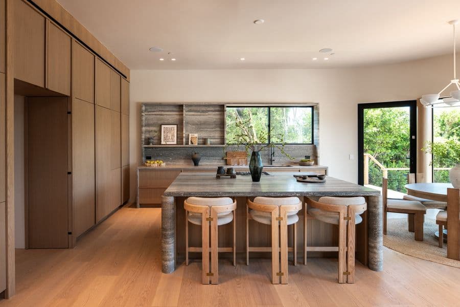 Kitchen with a full-height wall of rift white oak cabinets, a travertine island with upholstered stools, and a travertine backsplash under a wide window