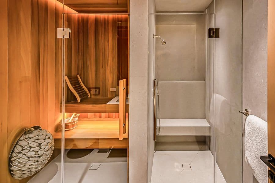 Custom clear cedar sauna with a slatted bench and backlit backrest, seen through a glass door beside a stone shower